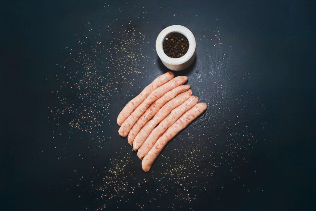 Raw chipolata sausages on a dark slate with a pot of peppercorns
